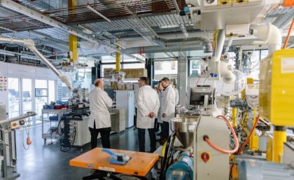 A group of people in white coats look at machinery inside a high-tech laboratory